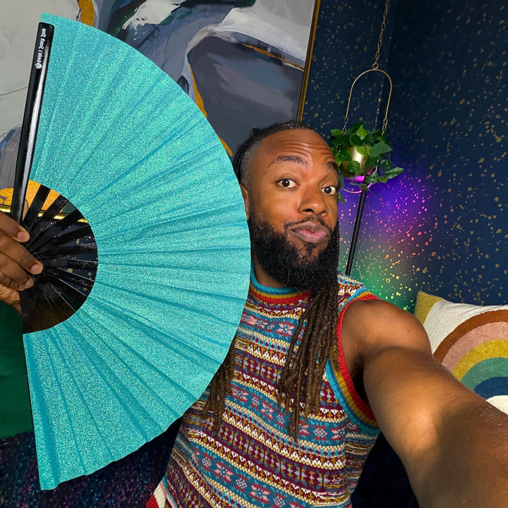 A man taking a selfie while holding up a glittery turquoise folding hand fan. He is wearing a colorful patterned sleeveless shirt, and the background features a dark blue starry wall, a hanging plant, and a rainbow-striped pillow.
