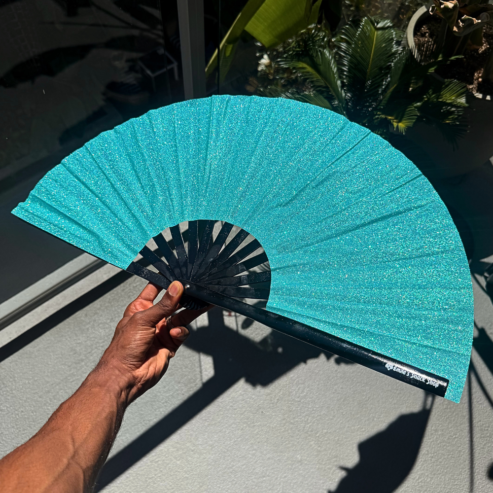 A hand holds a glittery turquoise folding hand fan with black ribs in front of a glass door with plants visible in the background.