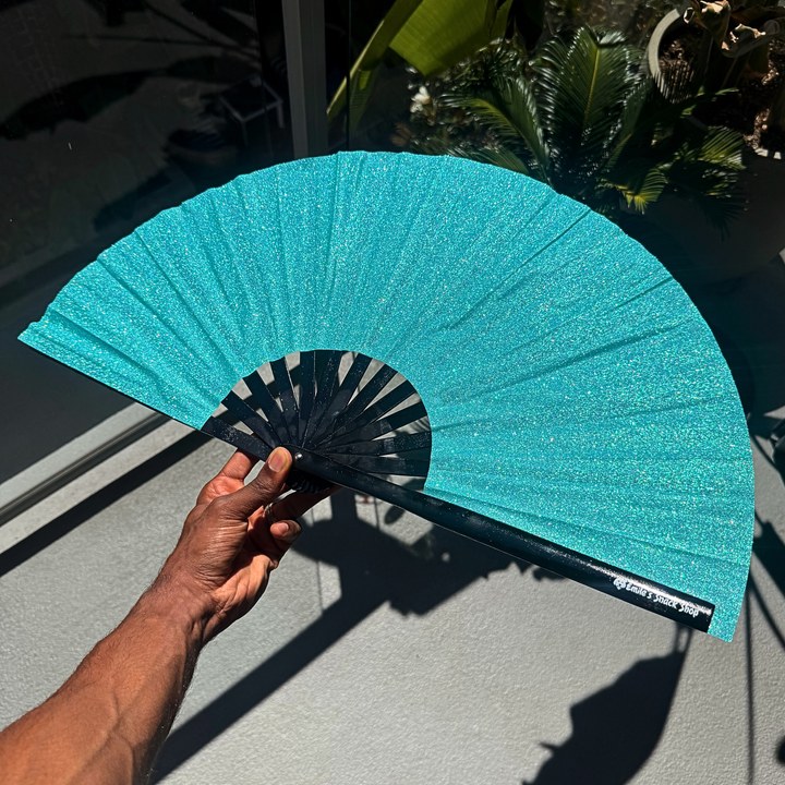 A hand holds a glittery turquoise folding hand fan with black ribs in front of a glass door with plants visible in the background.