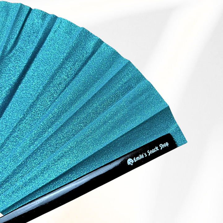 A close-up of a turquoise blue glitter folding fan with black ribs, showing the brand name &