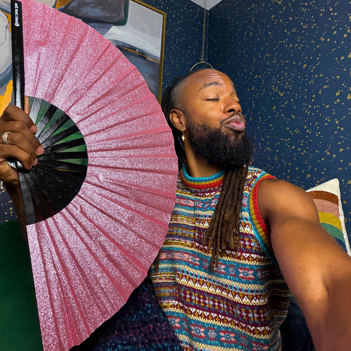 A man with a beard and dreadlocks poses in a colorful knit sweater while holding a pink glitter folding fan. The background features a dark blue wall with gold speckles.