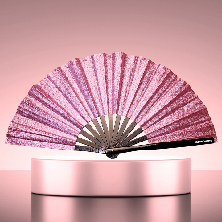 A pink glitter folding hand fan displayed open on a round pedestal against a soft pink studio background. The fan has a black handle with small white text near the end.