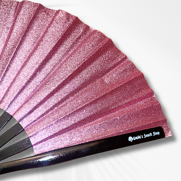 A close-up of a glittery pink folding hand fan with dark wooden ribs and a black label that reads &