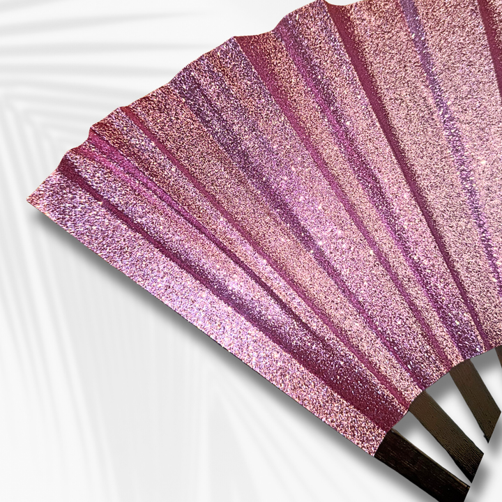 A close-up of a glittery folding hand fan with pink, purple, and rose gold shimmering stripes, shown against a white background with a subtle leaf shadow.