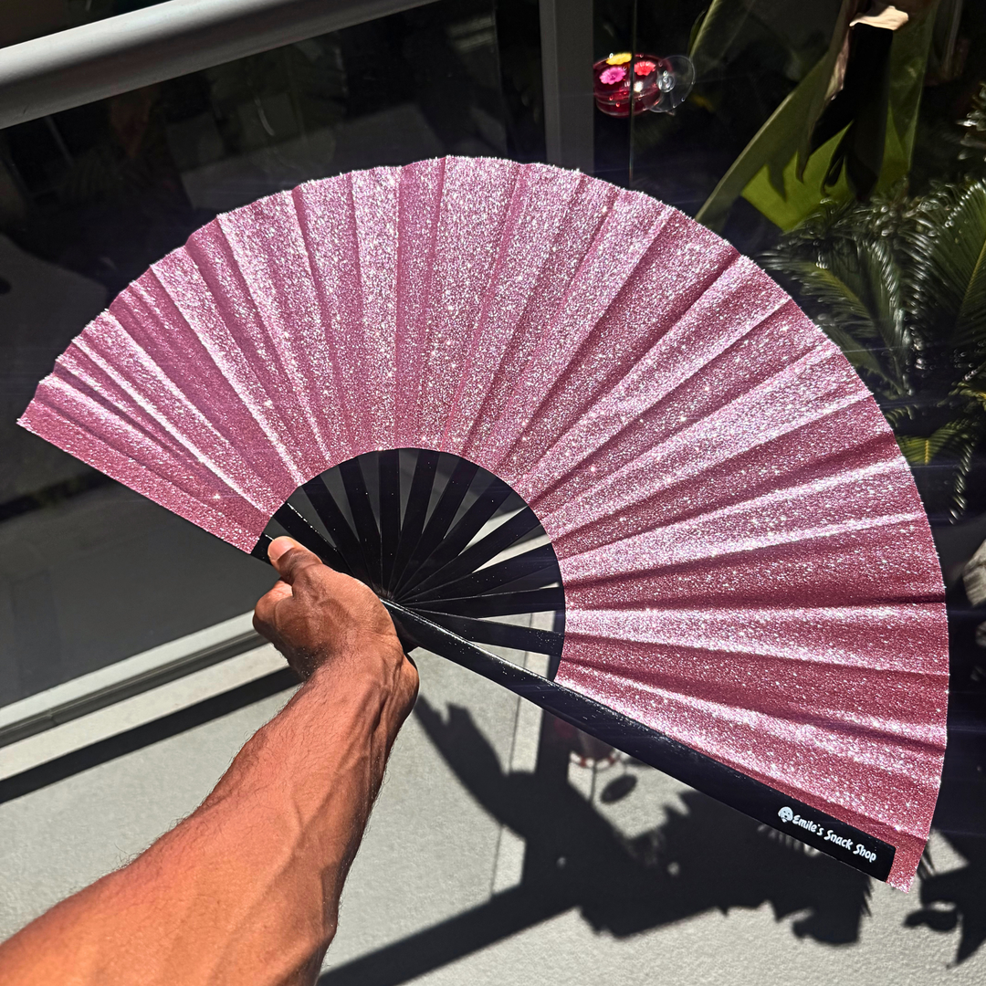 A hand holds a glittery pink folding hand fan with a black handle and ribs, photographed in bright sunlight near a glass door with reflections and shadows.