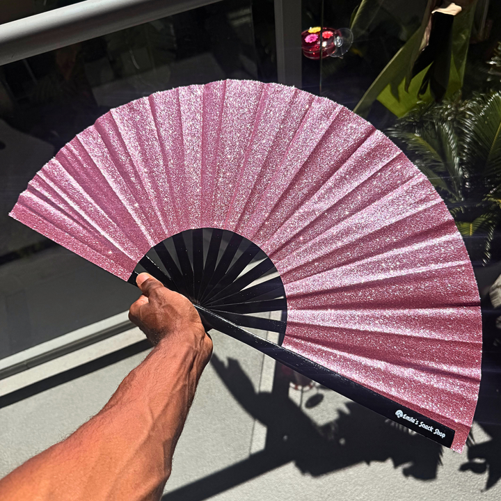 A hand holds a glittery pink folding hand fan with a black handle and ribs, photographed in bright sunlight near a glass door with reflections and shadows.