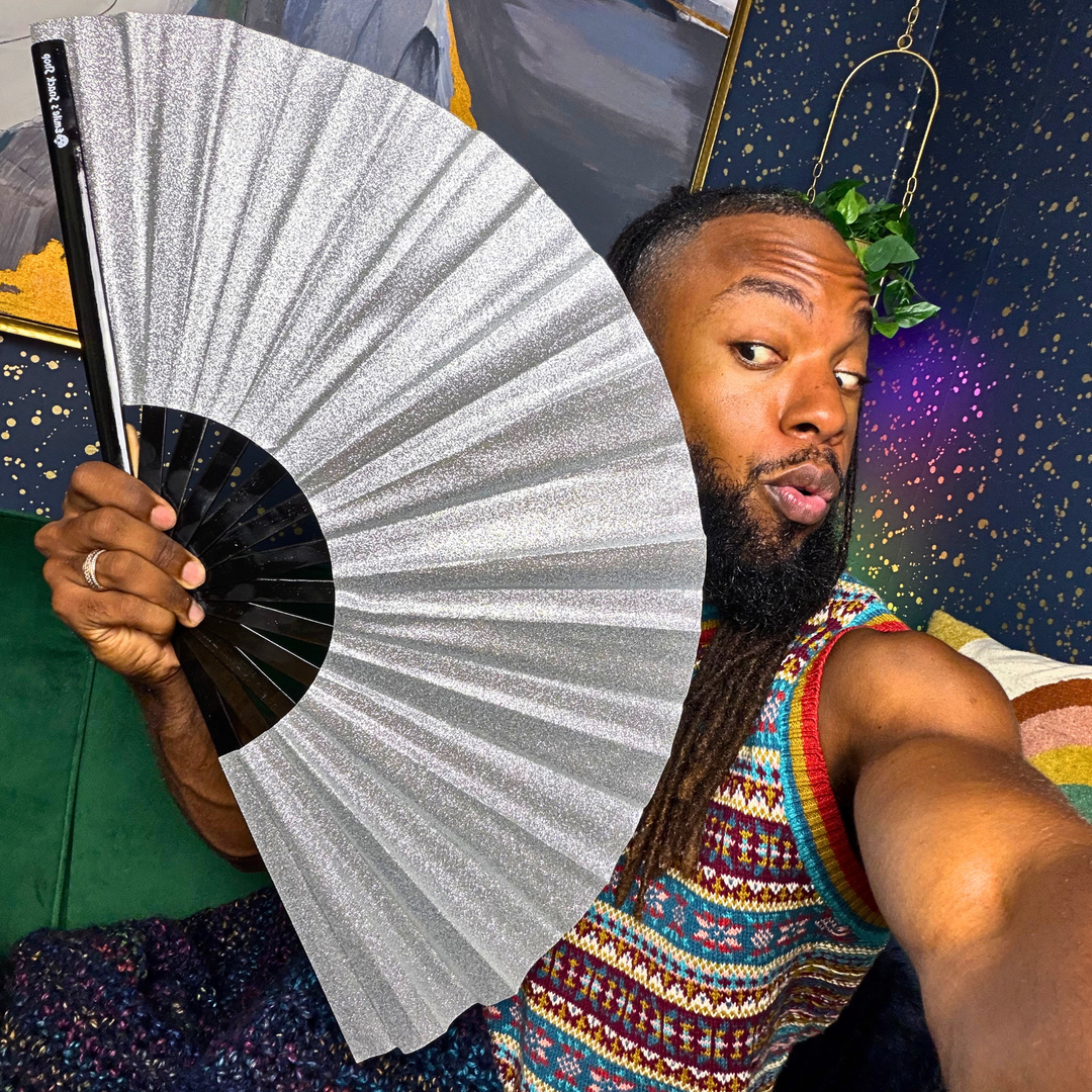 A man holding up a large silver glitter folding hand fan in front of his face, wearing a colorful patterned tank top and a green leaf headpiece in a decorated room.