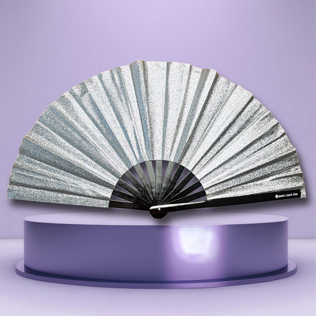 A silver glitter folding fan with black ribs, displayed open on a metallic purple pedestal against a lavender background.