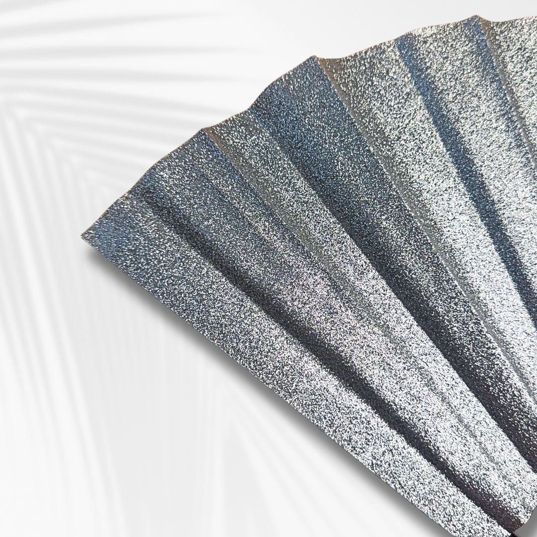 A close-up of a glittery silver folding fan with a textured, sparkly finish, shown partially unfolded against a clean white background with soft leaf shadows.