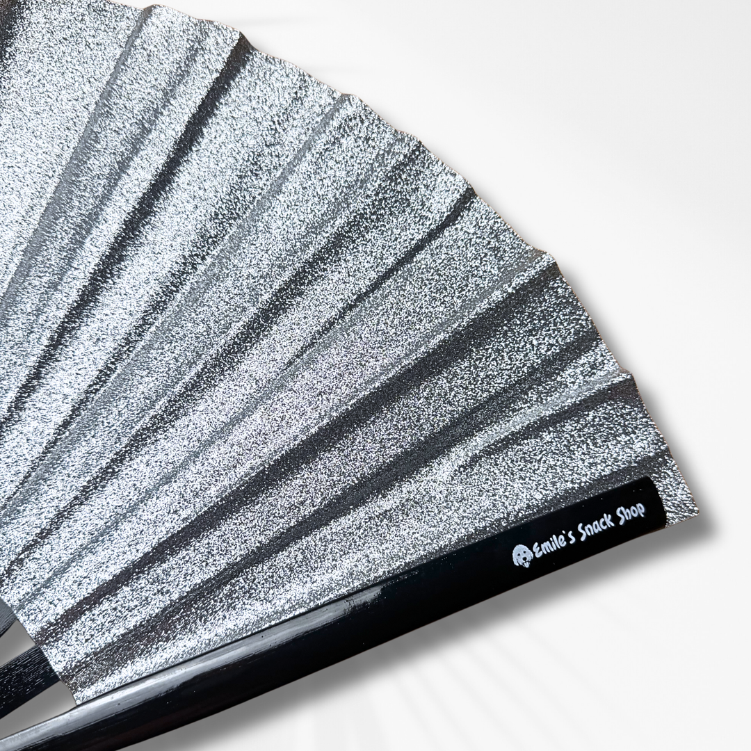 A close-up of a silver glitter folding hand fan with pleated ribs and a black handle, shown on a clean white background. The handle features a black label with the white text &