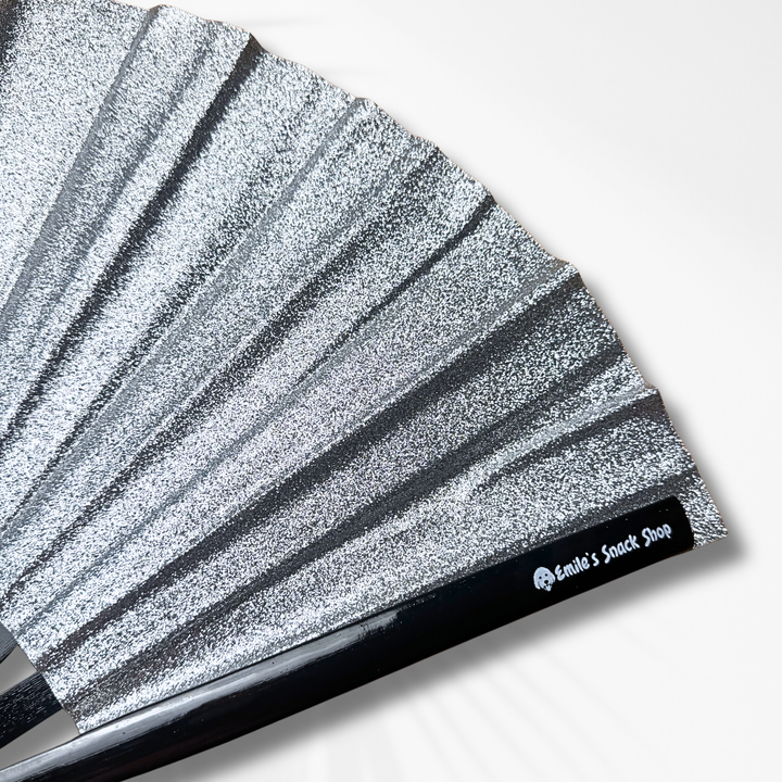 A close-up of a silver glitter folding hand fan with pleated ribs and a black handle, shown on a clean white background. The handle features a black label with the white text &