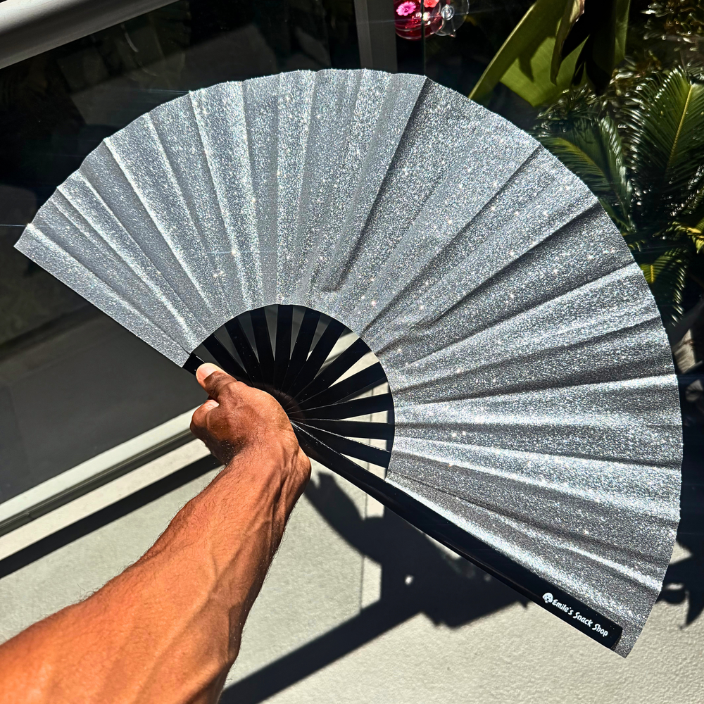 A hand holds a glittery silver folding hand fan with a black center and ribs, set against a background of a window and green plants.