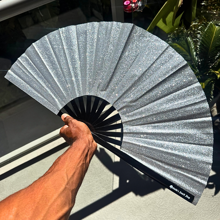 A hand holds a glittery silver folding hand fan with a black center and ribs, set against a background of a window and green plants.